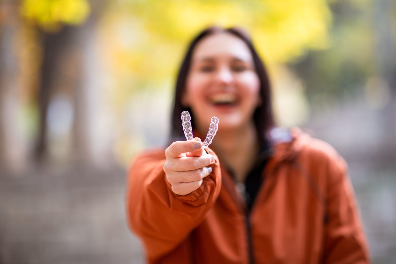 woman blurred out in the background in an orange shirt hold up her clear aligner. Invisalign Right for Me